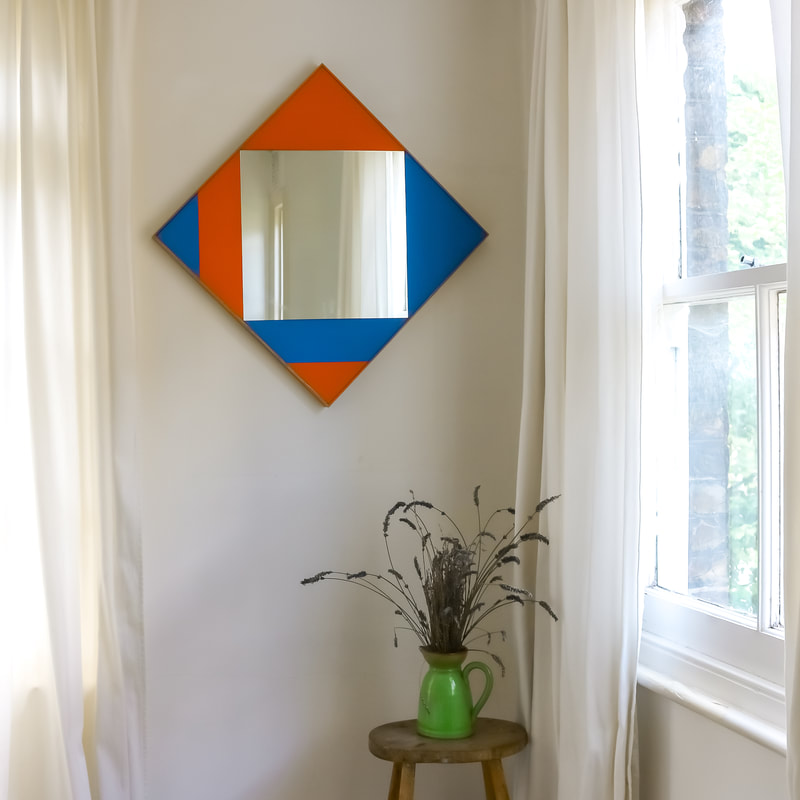 Square hand painted wall mirror in orange and blue in a white room
